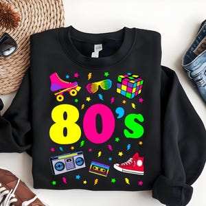 May include: Black sweatshirt with a colorful 80's theme design. The design includes roller skates, sunglasses, a Rubik's cube, a boombox, a cassette tape, and a pair of sneakers. The text "80's" is in large, colorful letters.