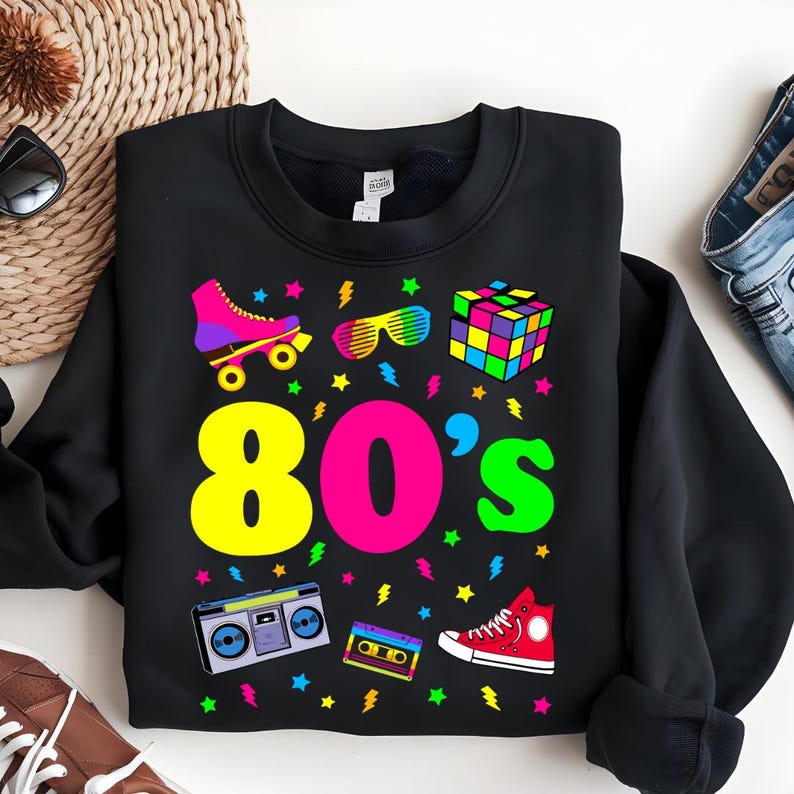 May include: Black sweatshirt with a colorful 80's theme design. The design includes roller skates, sunglasses, a Rubik's cube, a boombox, a cassette tape, and a pair of sneakers. The text "80's" is in large, colorful letters.