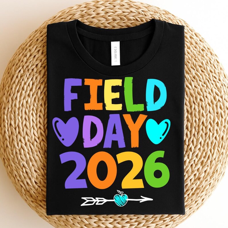 May include: Black t-shirt with the words "FIELD DAY 2026" in colorful letters. The design includes hearts, an arrow, and an apple. The shirt is displayed on a woven surface.