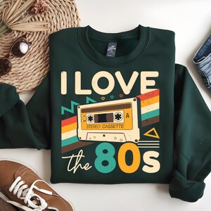 May include: A dark green sweatshirt with a retro-style graphic of a cassette tape and the text "I LOVE the 80s".