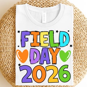 May include: White t-shirt with a colorful graphic that reads "FIELD DAY 2026". The text is in various colors, including yellow, green, blue, orange, and purple, with black outlines. The design includes hearts and stars.