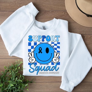 May include: White sweatshirt with a blue smiley face graphic. The smiley face has a blue ribbon and the text "Support Squad #Diabetes Awareness".