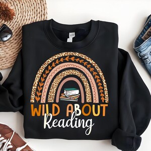 May include: Black sweatshirt with a rainbow design featuring a leopard print and a stack of books. The text "Wild About Reading" is printed below the rainbow.