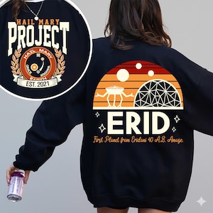 Puede incluir: Sudadera negra con un gráfico retro. La parte delantera presenta un diseño circular con el texto "HAIL MARY PROJECT" y "EST. 2021". La parte trasera muestra una escena de puesta de sol con la palabra "ERID" y el texto "First Planet from Eridani 40 A.B. Amage."