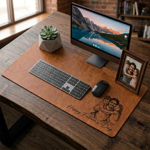 Könnte beinhalten: Eine braune Schreibtischunterlage aus Leder mit Tastatur, Maus und Computermonitor. Ein gerahmtes Foto und eine kleine Sukkulente in einem geometrischen Topf befinden sich ebenfalls auf dem Schreibtisch. Auf der Matte steht "Happy Father's Day".