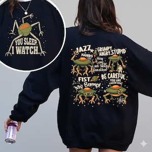 Puede incluir: Sudadera negra con un gran gráfico que presenta un diseño de robot verde y naranja y el texto "YOU SLEEP, I WATCH." La parte posterior de la sudadera tiene múltiples diseños de robots y frases como "JAZZ HANDS" y "BE CAREFUL."