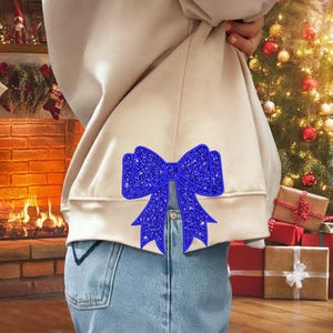May include: A beige sweatshirt with a large blue glitter bow on the back. The bow is centered on the back of the sweatshirt and is a festive holiday design.