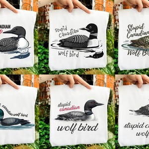 May include: White tote bags with a graphic of a loon swimming in water. The text "Stupid Canadian Wolf Bird" is above the illustration. The loon is black, white, and gray. The text is in a handwritten style font.