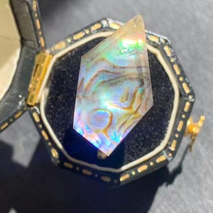 以下が含まれることがあります： 黒と金の六角形の宝石箱に入った、虹色のクリスタル宝石。 クリスタルは、青、緑、金色の渦巻き模様を持つプリズムの形をしています。 箱は黒いベルベットで裏打ちされています。