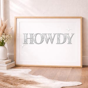 Könnte beinhalten: Gerahmter Druck mit dem Wort "HOWDY" in silbernen, umrandeten Buchstaben. Die Buchstaben haben eine strukturierte, glitzernde Optik. Der Druck befindet sich vor einem weißen Hintergrund und ist in einem hellen Holzrahmen gefasst. Dekorative Sternchen akzentuieren den Text.