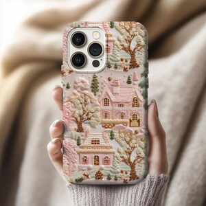 Puede incluir: Una funda de teléfono con un diseño en relieve 3D de un pueblo invernal. La funda es principalmente rosa y blanca, con casas en miniatura, árboles y nieve. La funda está sujeta en la mano de una persona.