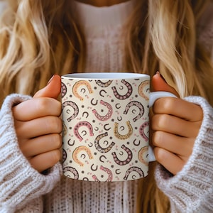 Puede incluir: Taza de cerámica blanca con un patrón repetido de herraduras en tonos marrón, rosa y amarillo sobre un fondo crema. Los diseños de herraduras están decorados con patrones florales y de puntos. La taza es sostenida por una persona que lleva un suéter blanco.
