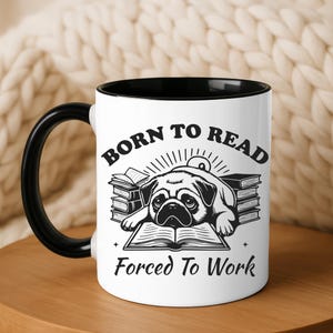 May include: White ceramic mug with a black handle and interior. The mug features a black and white illustration of a pug lying on books, with the text "BORN TO READ" above and "FORCED TO WORK" below the image.