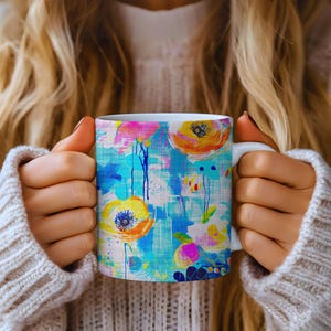 Puede incluir: Taza de cerámica blanca con un diseño floral colorido con flores abstractas amarillas, naranjas, rosas y azules. La taza tiene un asa blanca y un patrón vibrante y artístico, perfecto para disfrutar de su bebida favorita.