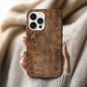 Puede incluir: Una funda de teléfono de cuero marrón con una superficie texturizada. La funda tiene un recorte para la cámara con múltiples lentes. La funda del teléfono está en la mano de una persona.