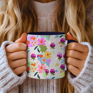 Puede incluir: Una taza de cerámica blanca con un estampado floral en rosa, morado, amarillo y verde. El interior de la taza es azul marino. La taza está sostenida por una persona que lleva un suéter blanco.