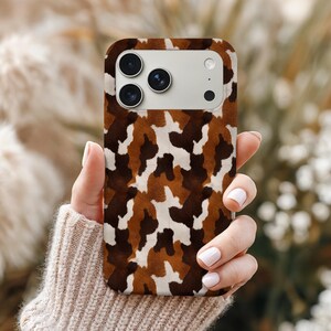 May include: A phone case with a brown, white, and tan camouflage pattern. The case has a rectangular shape with rounded corners and a camera cutout. The case is held in a person's hand, with a blurred background of greenery.