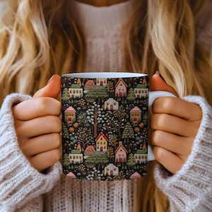 May include: White ceramic mug featuring a repeating pattern of colorful houses, trees, and foliage on a dark background. The houses have red, pink, and green roofs. The mug is held by a person wearing a cream-colored sweater.