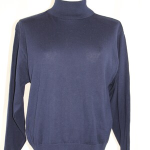 May include: A solid navy blue long-sleeve sweater featuring a high turtleneck collar, ribbed cuffs at the wrists, and a ribbed hemline at the bottom. The garment is displayed on a mannequin, showcasing a simple, plain knit construction throughout.