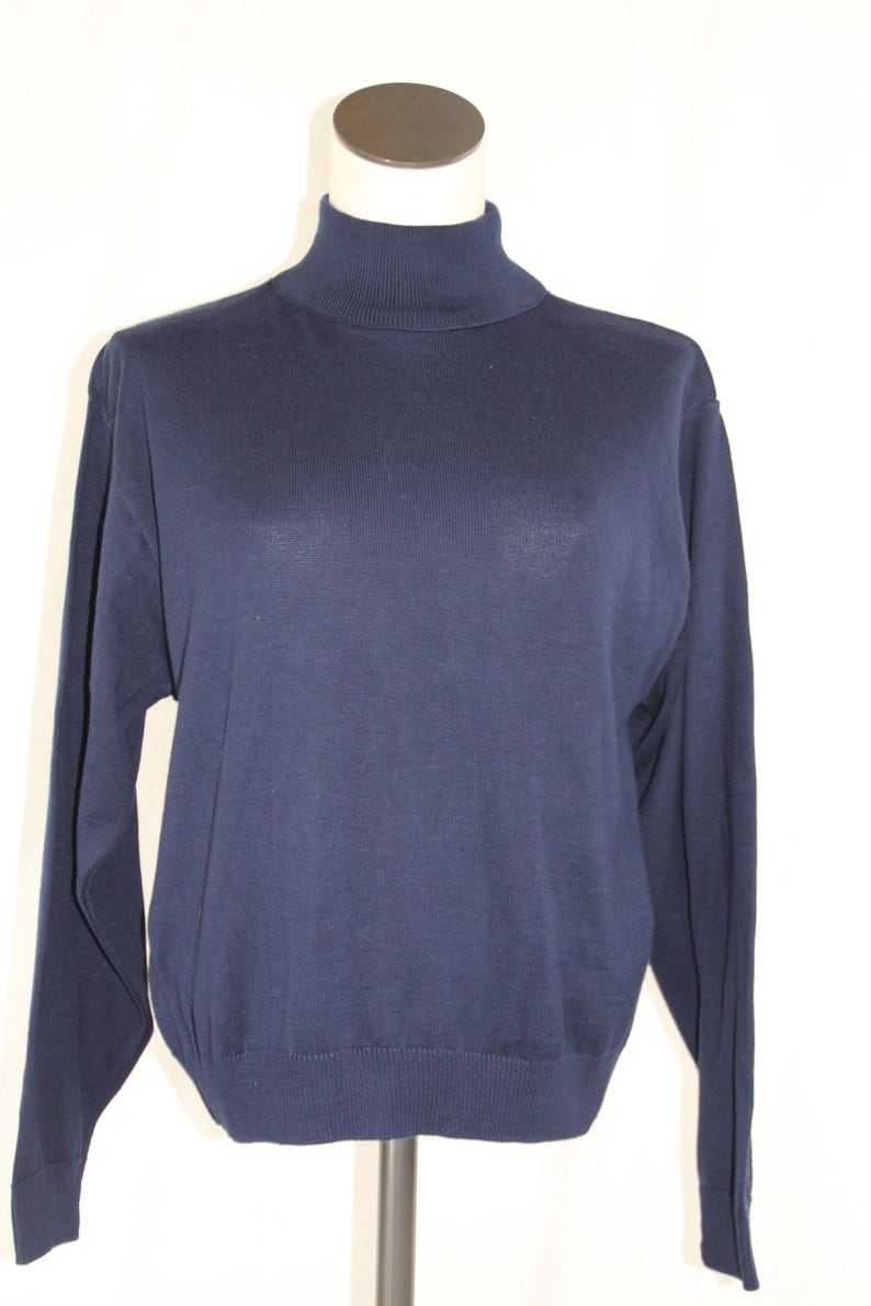 May include: A solid navy blue long-sleeve sweater featuring a high turtleneck collar, ribbed cuffs at the wrists, and a ribbed hemline at the bottom. The garment is displayed on a mannequin, showcasing a simple, plain knit construction throughout.