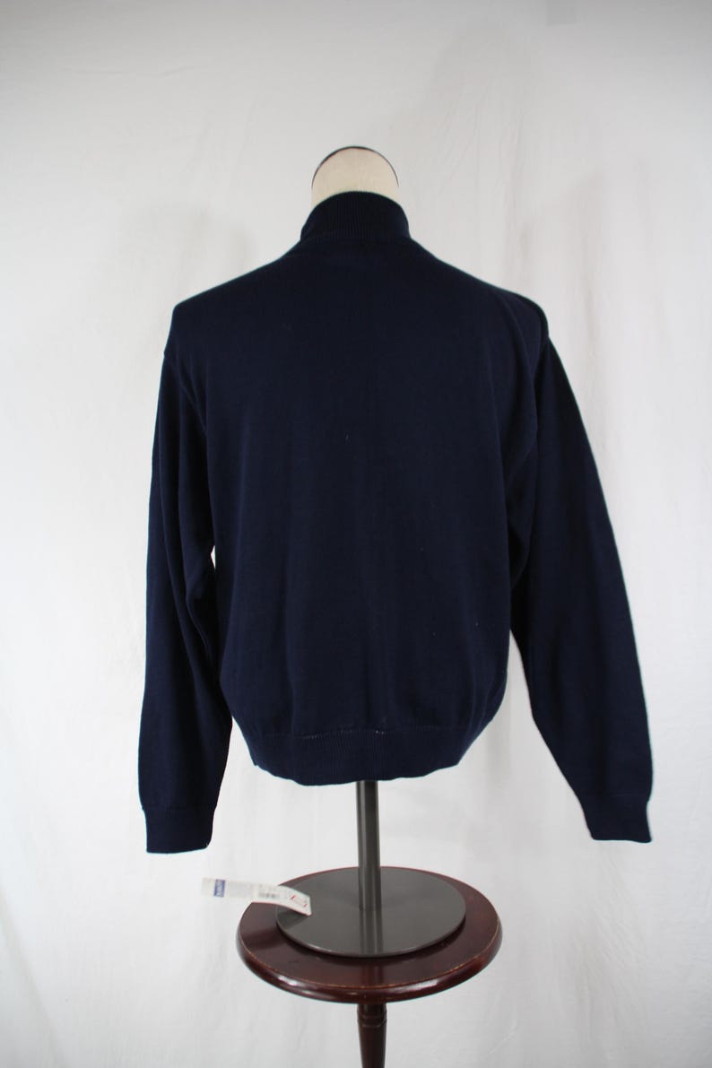 May include: A solid navy blue long-sleeve sweater featuring a mock neck collar, ribbed cuffs, and a ribbed hemline. The garment is displayed on a mannequin torso, showcasing a simple, plain back view of the knit pullover design.