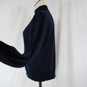 May include: A solid navy blue long-sleeve sweater featuring a mock neck collar. The garment is displayed on a mannequin, showing a side profile view with a fitted torso and sleeves that extend outward, highlighting the plain knit fabric construction.