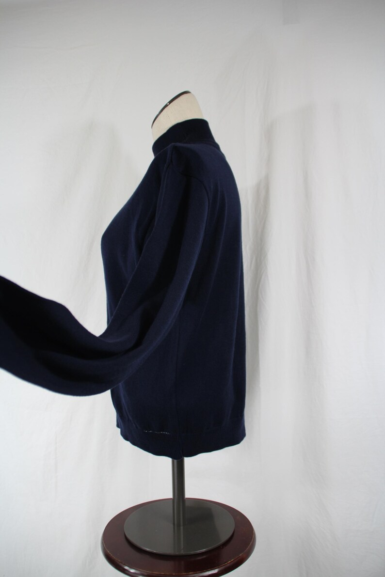 May include: A solid navy blue long-sleeve sweater featuring a mock neck collar. The garment is displayed on a mannequin, showing a side profile view with a fitted torso and sleeves that extend outward, highlighting the plain knit fabric construction.