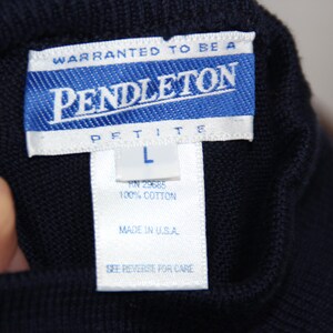 May include: A dark navy blue knit garment featuring a white rectangular tag with blue text that reads Warranted to be a Pendleton Petite L, 100% Cotton, Made in U.S.A., and See Reverse for Care. The fabric has a visible, uniform textured knit pattern.