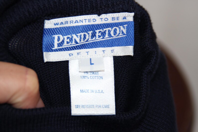 May include: A dark navy blue knit garment featuring a white rectangular tag with blue text that reads Warranted to be a Pendleton Petite L, 100% Cotton, Made in U.S.A., and See Reverse for Care. The fabric has a visible, uniform textured knit pattern.