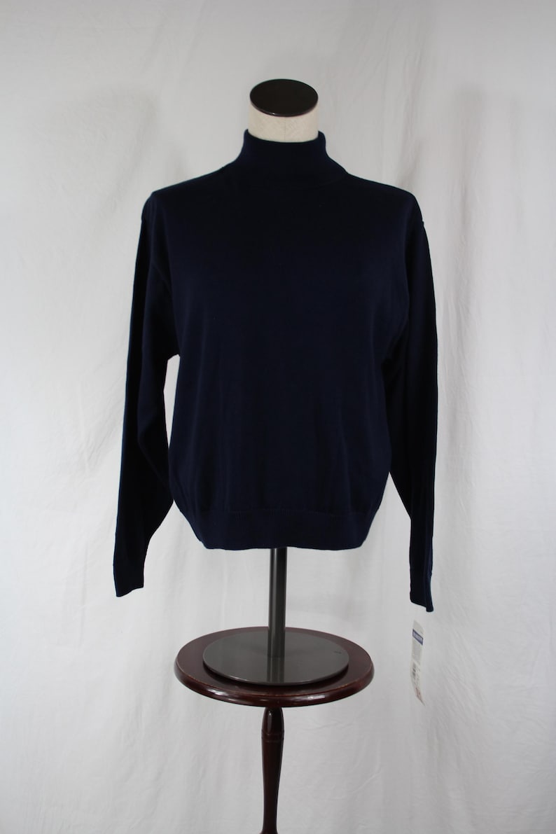 May include: A solid navy blue long-sleeve sweater featuring a high mock neck collar. The garment is displayed on a mannequin torso and includes a small white retail tag attached to the bottom hem on the right side of the piece.