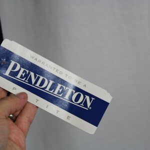 May include: A rectangular white and blue clothing tag with the text "WARRANTED TO BE A PENDLETON" in blue capital letters, with the word "PETITE" printed in smaller blue capital letters below the brand name on the bottom edge of the tag.