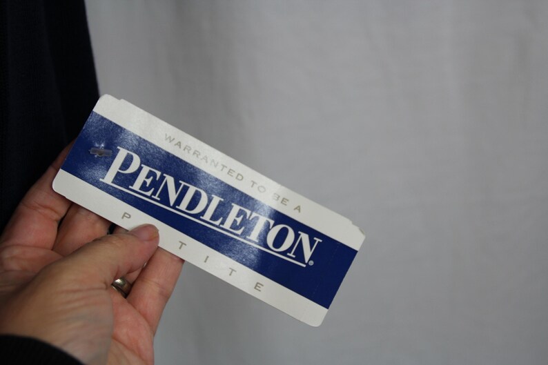 May include: A rectangular white and blue clothing tag with the text "WARRANTED TO BE A PENDLETON" in blue capital letters, with the word "PETITE" printed in smaller blue capital letters below the brand name on the bottom edge of the tag.