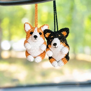 May include: Two crocheted Corgi ornaments hanging from strings. One Corgi is orange and white, while the other is black, tan, and white. Both feature small black eyes, stitched noses, and white chest fur, suspended by orange and black yarn loops.