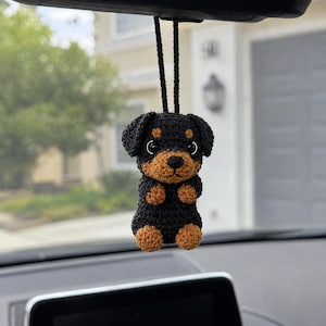May include: A small, handmade crochet Rottweiler puppy ornament hanging from a black cord. The figure features a black body with tan accents on the paws, muzzle, and above the eyes, complete with round black eyes and a small black stitched nose.