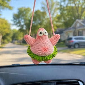 May include: A crocheted pink starfish ornament with two large white eyes and black pupils, wearing a ruffled green skirt. The figure is suspended by a pink yarn string, featuring a textured yarn construction with a small red stitched detail.
