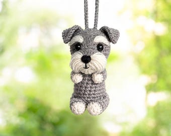 May include: A small, crocheted Schnauzer dog ornament in shades of grey and white. The figure features a grey body, white muzzle, black eyes, and a black nose, suspended by a matching grey crocheted loop for hanging as a decorative accessory.