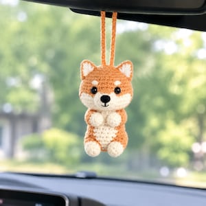 Könnte beinhalten: Ein gehäkelter Shiba Inu Hundeanhänger, der an einer Schlaufe hängt. Die Figur besteht aus orangefarbenem und cremefarbenem Garn, schwarzen Kunststoffaugen und einer gestickten schwarzen Nase. Sie hat spitze Ohren, kleine Pfoten und einen runden Körper.