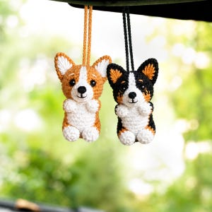May include: Two small crocheted corgi ornaments hanging from strings. One corgi is tan and white, while the other is black, tan, and white. Both feature pointed ears, dark eyes, and small paws, crafted with detailed yarn stitching and textures.