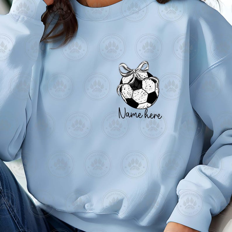 Puede incluir: Sudadera azul claro con un gr&aacute;fico de bal&oacute;n de f&uacute;tbol blanco y negro y el texto "Name here" impreso en la parte delantera.