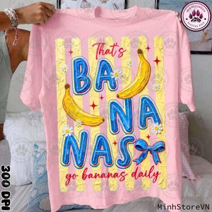 Puede incluir: Camiseta rosa con un gráfico colorido. El diseño presenta las palabras "That's BANANAS go bananas daily" en azul, plátanos amarillos y margaritas. La camiseta tiene cuello redondo y mangas cortas.