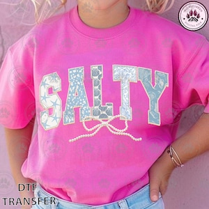 Puede incluir: Una sudadera rosa con la palabra "SALTY" en una fuente decorativa. Las letras están adornadas con patrones de conchas marinas y estrellas de mar, y debajo de la palabra hay un lazo de perlas. La sudadera es una transferencia DTF.