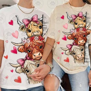 Puede incluir: Dos camisetas, una blanca y otra beige, cada una con un gr&aacute;fico de tres vacas de las Tierras Altas con lazos rosas, gafas en forma de coraz&oacute;n y coronas florales. Corazones rosas y rojos est&aacute;n esparcidos alrededor de las vacas. Las camisetas son usadas por dos personas.