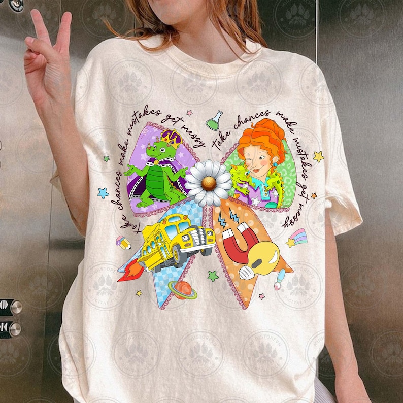 May include: Cream-colored t-shirt featuring a colorful graphic of a bow with cartoon characters, a school bus, and the text "take chances make mistakes get messy". The design includes stars and a rainbow.