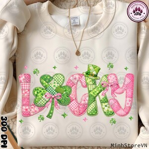 May include: Cream-colored sweatshirt with the word "Lucky" in patchwork style, featuring a green shamrock and a leprechaun hat. The letters are in pink and green, with a stitched design. The sweatshirt has a gold necklace.