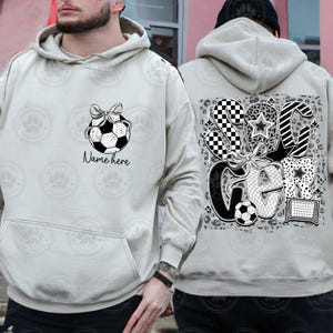 Puede incluir: Una sudadera con capucha gris claro con un gr&aacute;fico de bal&oacute;n de f&uacute;tbol blanco y negro en la parte delantera y un gr&aacute;fico de "Soccer" blanco y negro en la parte posterior. El texto "Name here" est&aacute; impreso debajo del gr&aacute;fico del bal&oacute;n de f&uacute;tbol.