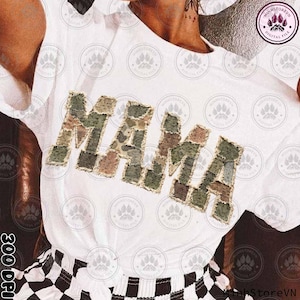 Può includere: T-shirt bianca con la scritta "MAMA" in un design patchwork mimetico. Le lettere sono in tonalità di verde, marrone e beige. La maglietta è indossata con pantaloncini a scacchi bianchi e neri.