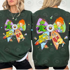 May include: Dark green sweatshirt featuring a colorful graphic. The design includes a bow with a daisy, a yellow school bus with flames, a dragon, and a woman with red hair. The sweatshirt has a white lining.