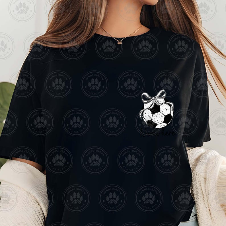 Puede incluir: Camiseta negra con un adorno de bal&oacute;n de f&uacute;tbol blanco con un lazo en la parte superior. El texto "Soccer Mom" est&aacute; debajo del adorno.