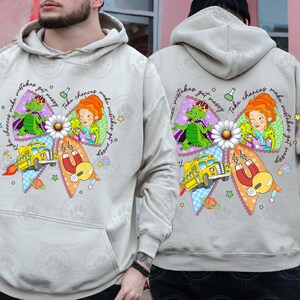 May include: A light gray hoodie featuring a colorful graphic design. The design includes a yellow school bus, a daisy, and cartoon characters. Text on the hoodie reads "Take chances make mistakes get messy."