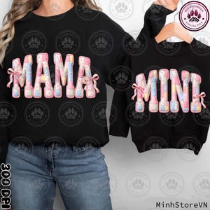 May include: Black sweatshirts with the words "MAMA" and "MINI" in a patchwork design. The letters are in pink, blue, and yellow with pink bows. The "MINI" sweatshirt is on a wooden hanger.